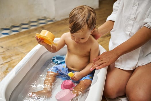 Adorable toddler enjoying bath time in a bathtub while playing with colorful toys. The child is supervised by an adult, creating a safe and fun bathing experience. Warm and caring atmosphere.