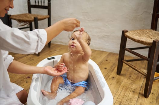 A cute baby in a small bathtub plays with water as a parent gently pours it. The setting is a warm and cozy room, creating a joyful and nurturing moment. Perfect for parenting themes.