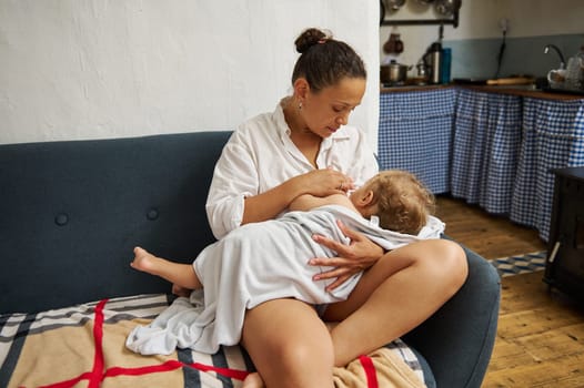 A mother sits on a comfortable sofa, tenderly breastfeeding her baby wrapped in a soft blanket. The cozy, homey environment highlights the bond and natural nurturing moment.