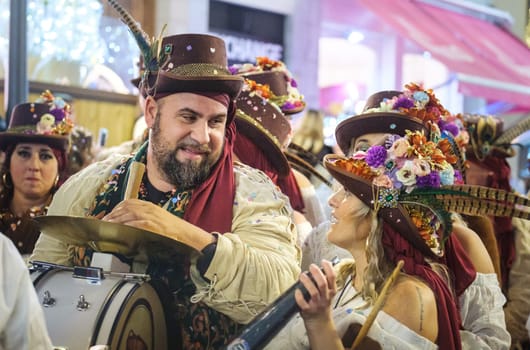 This image shows a group of people in festive attire that includes decorated hats with flowers and other adornments. A man, who is presumably a musician, is holding what looks like a drum, and he's sharing a joyful moment with a woman holding a wind instrument, possibly a clarinet. Both are smiling and seem to be engaged in a friendly interaction, indicating a lively and celebratory atmosphere, likely part of a festival or a traditional event. The background is a bit blurred, but it shows other individuals dressed similarly, suggesting a group performance or parade. The attire suggests that this could be a cultural or folk event. It's a colorful and vibrant scene.