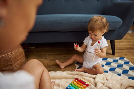 A curious toddler in a white onesie plays with a colorful xylophone in a cozy living room setting, exploring sounds and textures.
