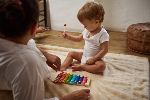 A mother and her toddler engage with a colorful xylophone on a soft carpet, encouraging learning and bonding through music in a cozy home setting.