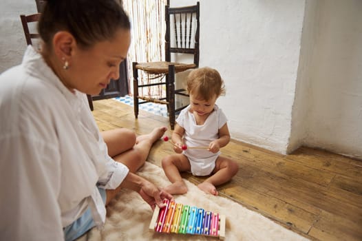 A mother and her young child sit on the floor, engaged in play with a colorful xylophone. The scene captures a warm, educational moment inside a cozy, rustic room, highlighting family bonding.