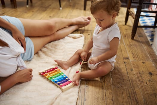 Cute toddler in a white onesie playing a colorful xylophone on a cozy blanket, next to a relaxed parent, creating a warm family atmosphere.