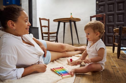 A happy mother and her baby enjoy bonding time playing with a colorful xylophone on a cozy wooden floor, showcasing joy and family connection.