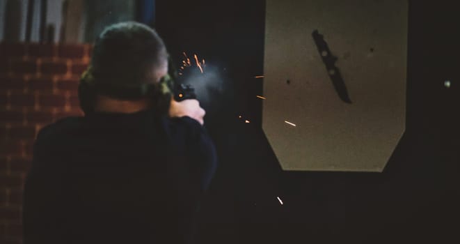 The image depicts a person at a shooting range. The person is seen from behind and is firing a handgun, with visible sparks and smoke coming from the gun. The target in front of them appears to be a paper or cardboard cut-out with a knife shape printed on it. The background includes a brick wall.