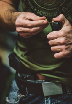 person loading bullets into a magazine, which likely belongs to a firearm. The person is wearing casual clothing, including a green shirt and jeans. visible holstered firearm at the person's waist.