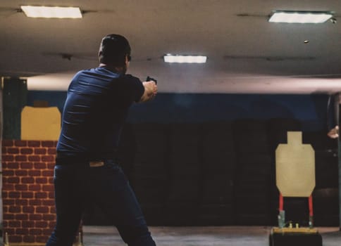 The image shows a person in a shooting range. The person is standing and aiming a handgun at a target downrange. The background includes typical shooting range elements such as an indoor setup with target silhouettes and protective barriers.