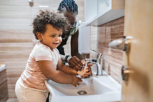 A playful toddler enjoys washing hands with guidance in a bathroom.