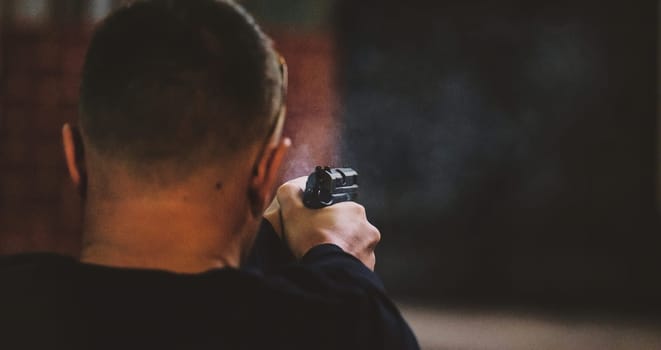 The image shows a person from the back aiming a handgun. The focus is on the firearm, which is visible in their hands, and the environment appears to be dimly lit. The context suggests a shooting range or similar setting.