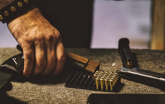 The image shows a person handling ammunition. There is a hand holding a handgun magazine, with a tray of bullets and parts of a firearm visible on a surface. The environment appears to be related to firearm handling or maintenance.