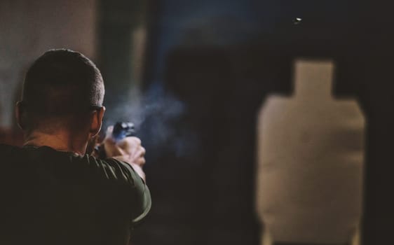The image shows a person shooting a firearm at a shooting range. The person is aiming at a target silhouette. There is visible smoke near the firearm, likely from a recent shot. The angle of the image is from behind the person, focusing on the act of shooting and the target ahead.