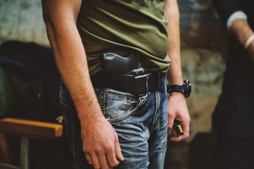The image shows the midsection of a person wearing jeans and a green shirt. The person has a handgun holstered at their waist. They are also wearing a black wristwatch on their left wrist. The background is somewhat blurred but appears to consist of a bench and possibly some other indistinguishable objects or people.