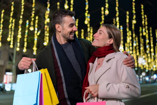 Smiling couple wearing Santa hats, standing under festive Christmas lights. They look at each other warmly, creating a joyful holiday atmosphere. One holds a phone, capturing memories.