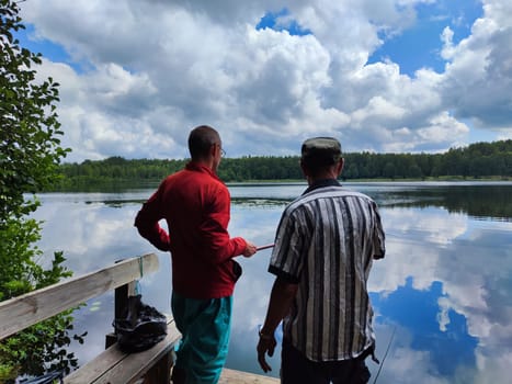 Two men converse and admire the calm water of the lake, surrounded by lush greenery and a dramatic sky scattered with clouds.