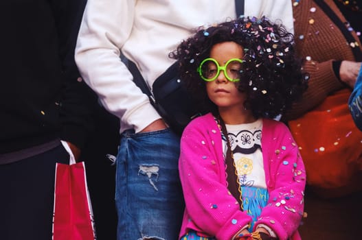 The image shows a young child with curly hair wearing large, round green sunglasses and a colorful pink cardigan over a white shirt with a character design. The child seems to be sitting down, surrounded by parts of two adults standing next to them