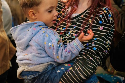The image shows a young child being held by an adult. The child appears to be looking with curiosity at something on their hand. Both the child's blue jacket and the adult's clothing are speckled with colorful confetti, suggesting they may be at a festive event, such as a party or celebration where confetti was thrown. The setting seems to be indoors with other people nearby, indicated by the partial view of another person in the background.