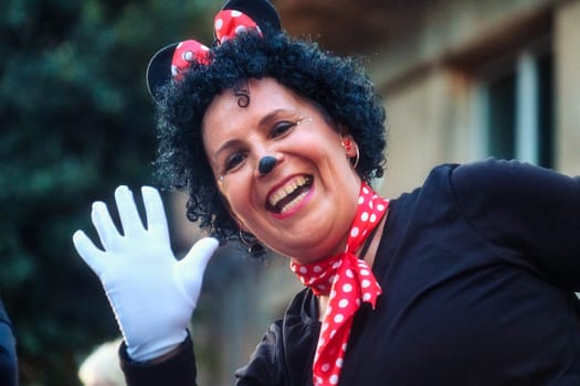 The image shows a person dressed in a costume resembling Minnie Mouse, a well-known animated character. The person is wearing a black top, a red headband with polka dots and mouse ears, and a red bow with white polka dots. They have face makeup that includes a black nose and they are smiling and waving to the camera, wearing a white glove on one hand much like the gloves often worn by various cartoon characters.