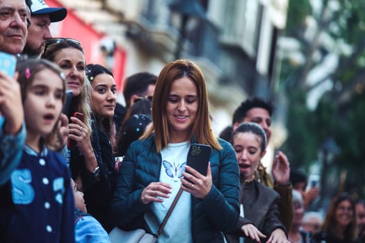In this image, there is a crowd of people, and they appear to be focused on an event or something of interest not shown in the photo. At the forefront, there is a woman with reddish-brown hair who is looking down at her smartphone, which she is holding with both hands. People in the background have various expressions, and there are a couple of individuals who are also capturing the moment with their phones. The setting looks like a city street with buildings in the background, suggesting an urban outdoor event or gathering. The shallow depth of field places the emphasis on the woman with the phone.