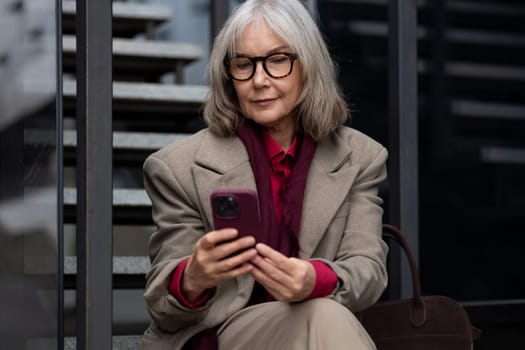 An elderly woman with gray hair is sitting on a set of steps outside, wearing a beige blazer over a red shirt. She is looking down at a smartphone in her hands.