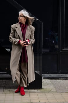 An elderly woman with gray hair wears a chic coat and red boots while standing against a dark glass building. The backdrop suggests a lively urban atmosphere in autumn.
