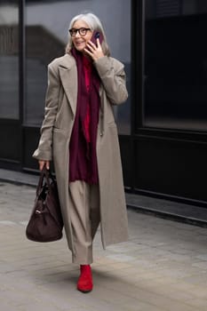 A senior businesswoman enjoys a stroll outside, talking on her phone. She wears a chic coat and vibrant red shoes, exuding elegance and confidence in an urban setting.
