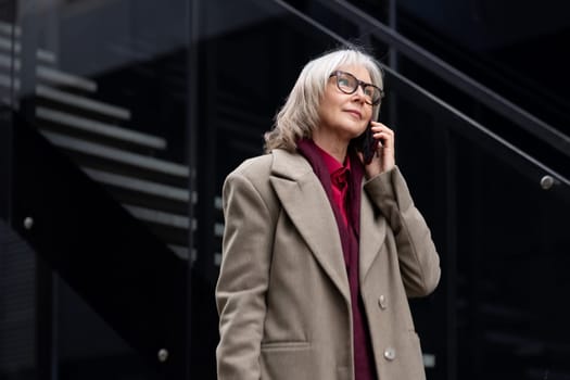 An elderly businesswoman wearing glasses and a stylish coat engages in a phone conversation outside a sleek office building, enjoying the warm weather.