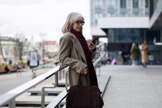 A senior businesswoman stands confidently outside a contemporary building, checking her smartphone while dressed in a stylish coat. The cool atmosphere suggests a brisk day.