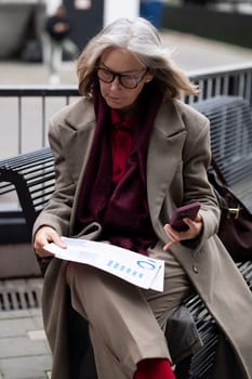 A thoughtful woman examines paperwork while seated on a bench in an urban setting. She holds a phone in her other hand and wears a stylish coat.