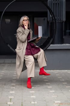 An elderly woman with a fashionable appearance sits thoughtfully in a circular outdoor seat, engaging with her laptop while enjoying the urban atmosphere on a sunny day.