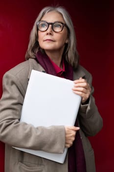 An elderly woman with stylish glasses and gray hair stands with a confident posture, holding a folder close to her chest against a vibrant red background.