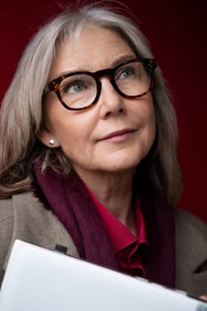 The older woman gazes upward with a pensive expression, wearing glasses and a scarf. She holds a notepad, suggesting contemplation or planning in a cozy setting.