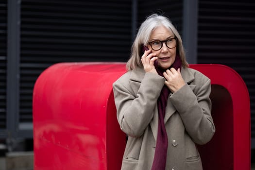 A middle-aged businesswoman with gray hair engages in a phone conversation while standing outside near a striking red structure on a gloomy day.