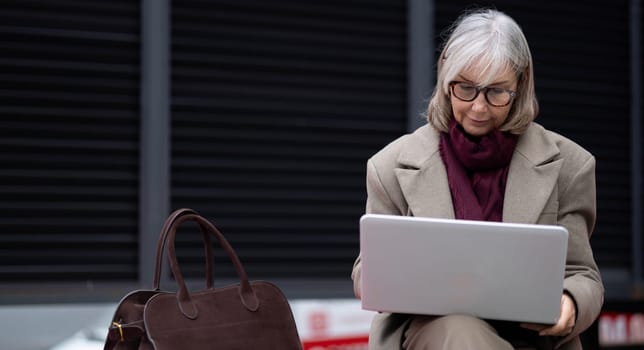 A senior businesswoman in a stylish coat works intently on her laptop outside, balancing her career with the flexibility of a city setting while seated with her bag.