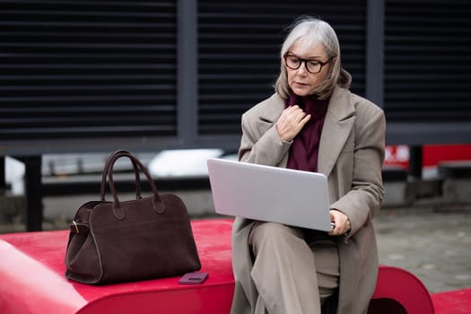 A senior businesswoman sits outdoors at a sleek bench, focused on her laptop. She wears stylish glasses and a light coat, with a handbag resting beside her, engaging in work.