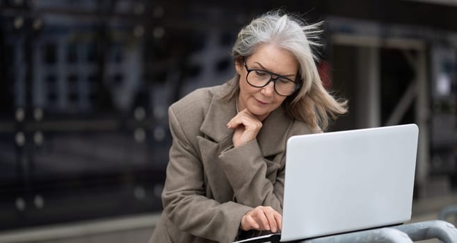 A mature woman with gray hair wears glasses as she focuses on her laptop, leaning thoughtfully in a bustling urban environment during a breezy afternoon.