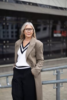 A fashionable elderly woman stands proudly by a railing, dressed in a stylish ensemble featuring a sweater with a V-neck design and a long coat, radiating confidence and elegance.