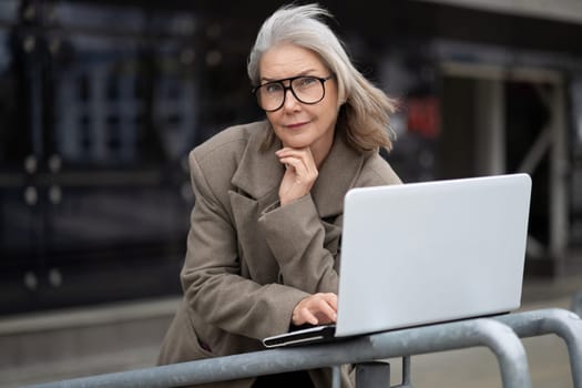 A middle-aged Caucasian businesswoman stands thoughtfully by a railing, typing on her laptop.