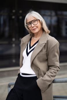 A senior businesswoman stands confidently outside a sleek modern building. Her glasses and stylish outfit highlight her professionalism as she poses casually.