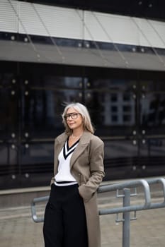 The senior businesswoman exudes confidence as she stands outside a sleek, contemporary building. Dressed in a chic outfit, she gazes thoughtfully, embodying professionalism.