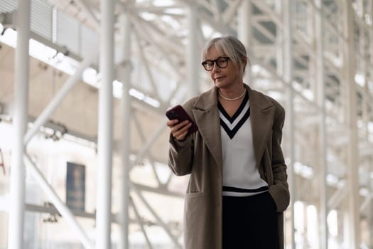 A middle-aged Caucasian businesswoman stands in a contemporary office space, absorbed in her smartphone, dressed elegantly in neutral tones against a sleek backdrop.