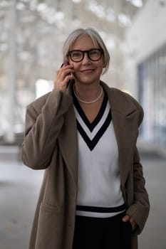 A confident elderly businesswoman wearing a stylish sweater and a coat stands in a contemporary office.