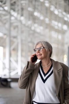 The elderly businesswoman, dressed elegantly in a sweater and blazer, smiles while conversing on her phone in a bright, contemporary office space, showcasing her confidence and poise.