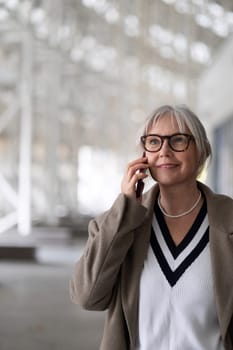 A senior businesswoman enjoys a phone conversation while strolling through a contemporary outdoor area. She appears confident and engaged, dressed smartly for the day.