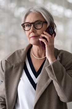 An older woman engages in a conversation on her phone, dressed elegantly in a coat and pearl necklace, expressing a moment of thoughtfulness and sophistication in her surroundings.