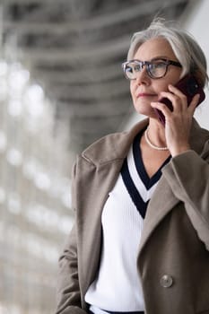A mature woman stands confidently, engaged in a phone conversation. She is dressed in a fashionable coat with a pearl necklace, surrounded by a contemporary architectural setting.