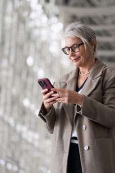 An elderly woman with grey hair and glasses smiles as she types on her smartphone, showcasing her stylish outfit in a modern indoor setting.
