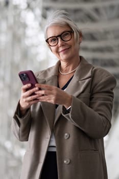 A senior businesswoman is smiling as she looks at her smartphone while standing outside in a contemporary urban environment during daylight.
