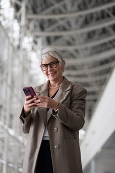 An older woman with silver hair and glasses stands indoors, smiling as she interacts with her smartphone, surrounded by a sleek, modern architecture.