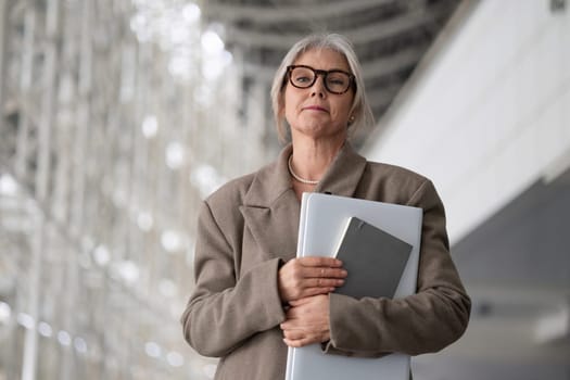 A middle-aged businesswoman stands confidently in a contemporary office setting, holding documents and a laptop. She appears focused and professional amid sleek architecture.